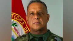 Javier Marcano Tábata ahagaze imbere y’ibendera. Yambaye impuzu za gisirikare, ariko araraba kamera.