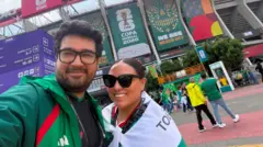 Homem e mulher tirando uma selfie em frente a um estádio de futebol, ele com uma jaqueta verde e ela usando uma bandeira verde e branca como capa