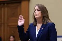 Kimberly Cheatle é uma mulher branca, de cabelos lisos e castanhos. Está vestida com um blazer azul marinho sobre camiseta branca, com um broche da bandeira dos Estados Unidos. Ela está com a mão direita erguida durante o juramento