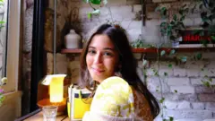 Mulher de cabelos castanhos e camiseta amarela sorri dentro de uma cafeteria com luz baixa.