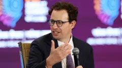 El director ejecutivo de Anthropic, Dario Amodei, se dirige a los asistentes en la Cumbre de Impacto de la IA, en Nueva Delhi, India. Viste gafas, un traje oscuro y una corbata morada, sentado frente a un fondo morado. Sostiene un micrófono mientras se dirige a alguien invisible.