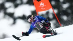 ヘルメットをかぶりゴーグルを着けた村岡が1本のスキー板にシート一体型のフレームを取り付けたチェアスキーで滑っている。両手には、先端が短いスキー板のようになったアウトリガーをもっている