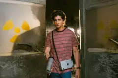 O ator Johnny Massaro como Márcio, de expressão séria, está à entrada de um ambiente industrial danificado, segurando um aparelho de medição com sensor manual e uma caixa presa ao corpo; nas paredes metálicas ao redor, há símbolos de radiação pintados.