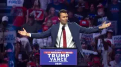 Uma imagem de Charlie Kirk discursando em um comício de Trump em Glendale, Arizona, em agosto de 2024. Atrás dele, uma multidão vestida com roupas vermelhas, brancas e azuis, segurando cartazes com os dizeres "VOCÊ ESTÁ DEMITIDO" e "LUTE! LUTE! LUTE!". Ele veste um terno azul com uma camisa branca de botões e uma gravata vermelha, discursando em um pódio enquanto gesticula amplamente com as mãos. No pódio, há uma placa com os dizeres "TRUMP-VANCE, FAÇAM A AMÉRICA GRANDE NOVAMENTE EM 2024".