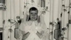 Foto preta e branca que mostra uma criança usando uma roupa branca, um véu e juntando as duas mãos em oração