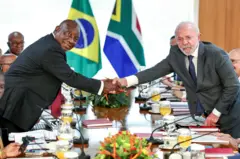 Imagem mostra os presidentes Cyril Ramaphosa, da África do Sul, e à direita, o presidente Lula. Os dois apertam as mãos sobre uma mesa de reuniões.