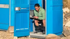 A man sitting on the toilet in a portable toilet in the middle of the Death Valley desert, in the United States. He has his trousers rolled around his ankles and is reading his phone. The toilet door is wedged open by a rock and another portable toilet is next to hium, with the door closed. Rocks are scattered around and there are barren hills in the background.