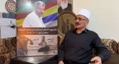 رجل مسن يجلس وبجانبه صورة شقيقه المقتول