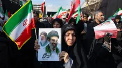 Mulher em Teerã vestida com um xador preto, segura uma bandeira do Irã e um cartaz com uma imagem do novo líder supremo do país, Mojtaba Khamenei, ao lado do seu pai, o aiatolá Ali Khamenei. Ela está em uma multidão de pessoas com objetos similares.