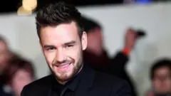 Liam Payne, wear black shirt, suit and tie, smile for photographers for one red carpet event wit several onlookers behind am