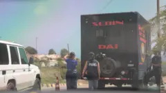 Caminhão do Sebin, dois agentes e uma jornalista com jaleco dizendo 'imprensa' em espanhol, em La Guaira, Venezuela, julho de 2026