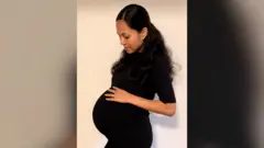 Jenitar Na'amoana fotografada grávida. Ela tem cabelos longos e escuros e veste um vestido preto de mangas curtas. Ela está olhando para baixo e tocando sua barriga de grávida bem visível.