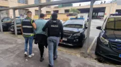 Un hombre con camiseta azul de manga larga flanqueado por dos agentes de Policía protegidos por chalecos balísticos. Caminan hacia autos policiales estacionados. 