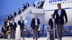 飛行機から降りて来たダークジャケットにグレーパンツ姿の選手らが荷物などを手に滑走路を歩いている
