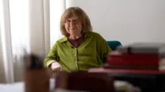 Світлана Алексієвич сидить за столом на тлі білої стіни і заштореного вікна в оливковій сорочці. На передньому плані олівці і ручки в підставці і корінці книг.