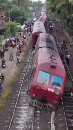 දුම්රිය එන්ජිමක් සහ මැදිරි කිහිපයක් පීලි පන ඇති අතර දුම්රිය මග දෙපස මිනිසුන් සිටින අයුරු පෙන්.