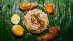 Ninu ewe ọgẹdẹ to tutu dódó, wọn bu irẹsi si aarin ewe naa, ẹyin aabọ ati odidi kan wa lẹgbẹẹ rẹ, bẹẹ ni awọn ẹran yi i ka pẹlu. Alubọsa ti wọn rẹ si i lori wa nibẹ naa, wọn si fi ata ṣọmbọ kan ti ko ti i pọn le ori irẹsi ti wọn bu ata dindin le lori naa pẹlu. 