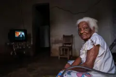 Na foto está Eulalia, uma mulher idosa com mais de 90 anos. Ela é negra, tem cabelos brancos e está sentada em uma cadeira. Na frente dela há uma televisão