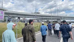 Багато людей рухаються пішки вздовж дороги під естакадою, частина з них зупинилася і стоїть уздовж огородженого тротуару.
Над ними — широкий міст із ліхтарями, а на тлі видно хмарне небо.
