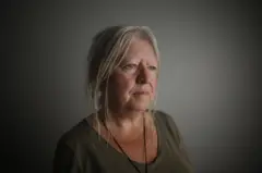 Una mujer con cabello rubio canoso hasta los hombros mira hacia un lado. Está de pie frente a una pared clara. Lleva una blusa color oliva.