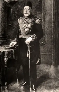 Mehmed V Reshad, de barba blanca, está parado al lado de una mesa. Una de sus manos reposa sobre unos libros y la otra sostiene lo que parece ser una espada que toca el suelo. Viste un elegante traje militar y lleva puesta un tarboosh