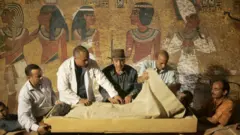 Con la ayuda de técnicos en mangas de camisa y bata de laboratorio, el jefe de antigüedades de Egipto, Zahi Hawass (centro), con un sombrero fedora marrón al estilo de Indiana Jones, supervisa la extracción de la momia de Tutankamón, envuelta en lino, de su sarcófago de piedra, en su tumba subterránea, en el Valle de los Reyes, en Luxor, el 4 de noviembre de 2007. Arte egipcio antiguo, que muestra figuras con cabezas y piernas de perfil pero torsos de frente, y jeroglíficos adornan la pared detrás.