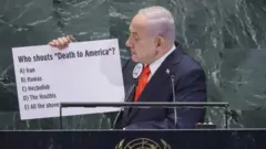 Netanyahu exibindo um cartaz em que pergunta "Quem grita morte à América?", seguido de opções que incluía os Houthis, o Hezbollah, o Hamas, o Irã ou "todas as opções acima".