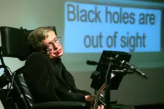 Stephen Hawking em sua cadeira de rodas, usando uma roupa preta. Atrás dele há um painel onde se lê: Buracos negros estão fora nosso campo de visão.