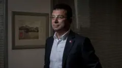 Ekrem İmamoğlu üzerinde bir takım elbiseyle, kravatı olmadan loş bir ofiste kadrajın solundan uzaklara bakıyor