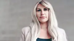 Rebecca posa diante de um muro de pedra clara. Ela tem cabelos loiros longos e ondulados, veste uma blusa preta e uma jaqueta rosa-clara e olha para a câmera com expressão pensativa
