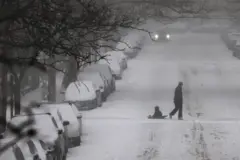 شخص يتنقل وسط الثلوج في شارع تكسوه الثلوج بالكامل وتصطف السيارات على الجانبين مغطاة كاملةً بالثلوج.