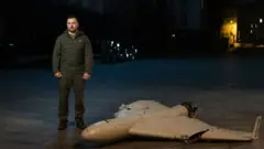 ዘለንስኪ የተመታች ድሮን ጎን ቆመው 