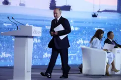 Володимир Путін у чорному костюмі з папкою в руках йде по сцені до білої трибуни на фоні величезного екрана з зображенням кораблів