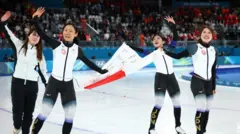 白と黒のウエアを着た4人が笑顔で、日本国旗を持ちながらリンクで滑っている。佐藤以外の3人は片手を上げている