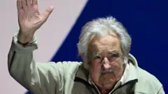 José "Pepe" Mujica saluda con una bandera de su partido, el Frente Amplio, detrás.