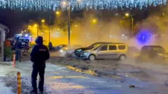 Прорыв трубы в Санкт-Петербурге в декабре 2025 года: ночная улица с новогодними украшениями на столбах и горящими фонарями, спецтехника на заднем плане. В воздухе пар