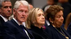 Bill ve Hillary Clinton'ın 14 Mayıs 2025'te katıldıkları bir cenaze töreninde çekilmiş bir fotoğrafı. Yan yana oturuyorlar, Bill Clinton'ın yakın gözlükleri var ve biraz şaşırmış bakıyor. 