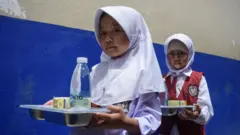 Dua murid membawa nampan dengan menu MBG di sebuah sekolah dasar di Sumedang, Jawa Barat, pada 18 November 2024.
