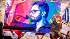 মঞ্চে উমর খালিদ (পর্দায়) দবং আরও যারা বন্দী হয়ে আছেন, তাদের জন্য ন্যায়বিচারের দাবী তোলেন শিল্পী রূপম ইসলাম (সামনে)