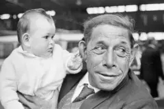 A baby in a white cardigan gently touches the ear of the man holding him. The man, dressed in a jacket and tie, looks away from the child. Both are among the Tristan da Cunha islanders newly arrived in the UK following the 1961 volcanic evacuation