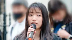 Chloe Cheung speaking into a microphone at a protest in the UK, with two young protesters standing behind her, whose identity is hidden by a blurring technique 