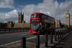Ônibus vermelho de dois andares passando pela rua em Londres. No fundo está o Big Ben e o parlamento britânico