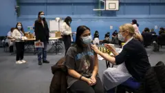 Bir kadın öğrenci kolunu açmış bir kadın hemşire ona aşı yapıyor. Kapalı spor salonu gibi bir alanda küçük masalarda çok sayıda aşı yapılıyor. İki kadın öğrenci ayakta sıra bekliyor. 