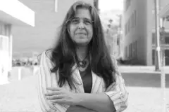 Carla Molento, uma mulher de meia idade de cabelos longos grisalhos e óculos, de braços cruzados olhando para a câmera. Ela veste um terno feminino listrado e usa um colar com formato de borboleta