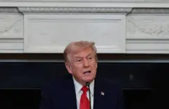 Trump falando ao microfone em salão interno, com feição séria