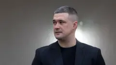 Портретне фото чоловіка в темному піджаку та чорній футболці на нейтральному розмитому тлі. Людина з короткою стрижкою дивиться вбік, знімок зроблено в приміщенні під час офіційного заходу або публічного виступу.