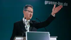 Mikrofon başında gözlüklü bir adam gülümseyerek eliyle kalabalığa işaret ediyor. Arkada Yeşiller Partisi'nin logosu var. 