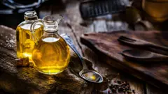 Two bottles of oil and one spoil with oil in it. Other kitchen utensils are in the frame.