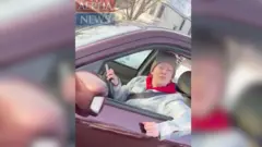 赤紫色のSUVの運転席に、グレーのニット帽をかぶり、白と水色のジャケットに赤いフード付きパーカーを着た女性が座っている。女性は左腕を窓の外に出し、右手をハンドルに乗せ、ほほえみながらカメラの方を見ている