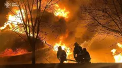 Троє рятувальників у захисному спорядженні, які стоять на колінах і тримають пожежний шланг, спрямований на величезне полум’я. Вогонь охопив велику конструкцію на задньому плані, полум’я яскраво жовтогаряче, з густим димом. Попереду – темний схил із кількома голими деревами, що створюють силуети на тлі пожежі.